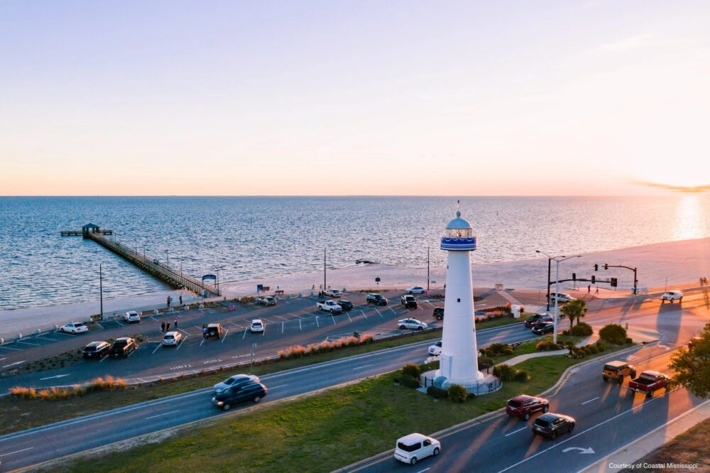 White lighthouse on grassy point overlooking Gulf of Mexico beach with pier, parked cars, and sunset sky