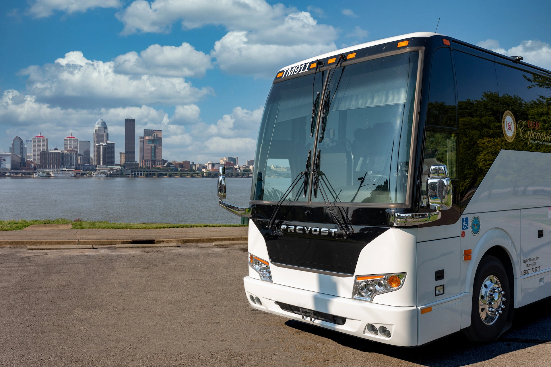 White and black charter bus parked at riverfront with Louisville skyline and Ohio River in background under blue sky