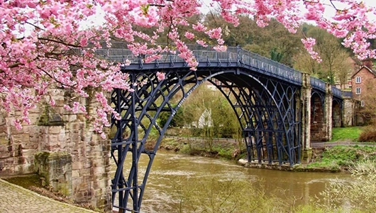 Cast iron footbridge spanning calm waterway with blooming pink cherry blossom trees and historic stone buildings